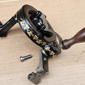 May include: Black antique sewing machine hand crank with gold floral accents and a wooden handle. A silver screw is next to the crank. The crank is on a light brown surface.
