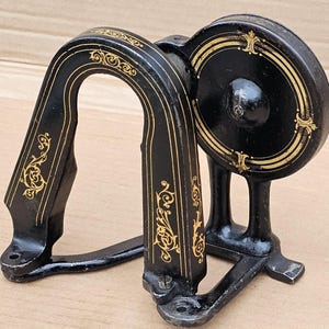 May include: Antique black sewing machine component with gold filigree detailing. The component features a curved handle and a circular wheel, both adorned with ornate patterns. A vintage piece.