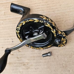 May include: A vintage black sewing machine hand crank with gold floral detailing. The crank features a black wooden handle and a silver-colored metal arm. A small screw is also present.