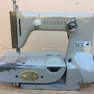 Op de afbeelding: Een grijze Pegasus DM-10 naaimachine met een metalen voetpedaal en een label met de tekst "Hyde & Wakely, Machinists, Electricians, The Triangle, Yeovil".