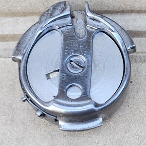 May include: A silver-colored sewing machine bobbin case. The case has a circular shape with a central metal piece secured by a screw. The text "ENGLAND" and "6 - 12" are visible. The case has a curved top and a small hook.