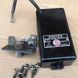 Puede incluir: Un controlador de motor de costura Singer negro con un cable negro y un soporte metálico. El controlador tiene una etiqueta con texto y el logotipo de Singer. También hay varios tornillos presentes.