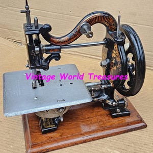 May include: Antique black sewing machine with ornate gold detailing and a wooden base. The machine features a hand-crank wheel and a needle, showcasing a vintage design. The text "Vintage World Treasures" is visible on the image.