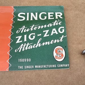 Puede incluir: Folleto Singer Automatic ZIG-ZAG Attachment vintage con un tornillo de metal plateado. El folleto es verde y naranja con texto blanco, incluyendo "SINGER" y "Automatic ZIG-ZAG Attachment". También es visible el logotipo de Singer Sewing Machines.