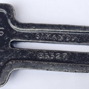 May include: A vintage, dark gray metal tool with a distinctive shape. The tool has a rectangular body with a curved end and a central elongated opening. The text "SIMANCO CO." and the number "85527" are visible.