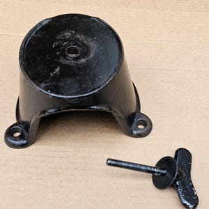 May include: A black, cast iron object with a circular top and a flared, curved base. Two holes are visible on the base. A threaded bolt with a wing nut is next to the object. The items appear to be vintage.