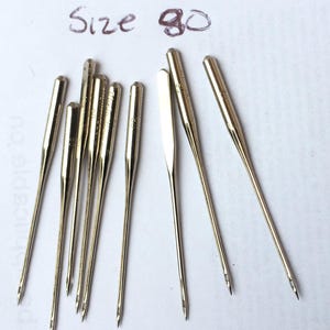 Puede incluir: Un conjunto de agujas de máquina de coser doradas. El texto manuscrito "Size 90" en tinta morada está encima de las agujas. Las agujas están dispuestas sobre una superficie blanca, con las puntas hacia abajo.