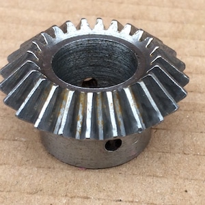 May include: A silver metal gear with a large central hole and a smaller hole on the side. The gear has a series of teeth around the outer edge.