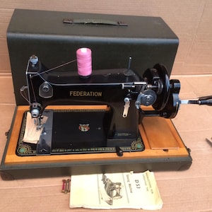 May include: A vintage black Federation sewing machine with a wooden base and a green carrying case. The machine has a pink spool of thread and the word "FEDERATION" is printed on the front. The machine also has the word "BRITISH" printed on it.