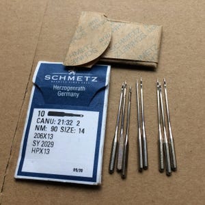 Puede incluir: Un juego de agujas para máquina de coser Schmetz, tamaño 14, con embalaje. El embalaje azul y blanco indica que las agujas son de Herzogenrath, Alemania. Se ven varias agujas plateadas.