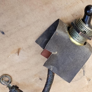May include: Close-up of an electrical switch with a black knob and metal components. The switch is partially covered by a dark gray, textured material. A section of a wire is visible, along with a metal ring.