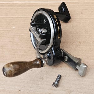 May include: A vintage sewing machine hand crank with a wooden handle and a black metal frame. The crank features a silver-colored wheel and a detached screw. The design includes an arrow detail.