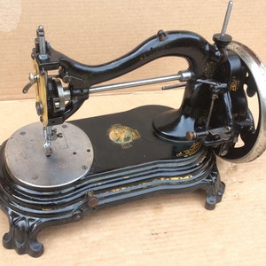 May include: A black vintage sewing machine with a metal foot pedal and a decorative base. The machine has a gold-colored trim and a small, round, silver-colored plate on the front.