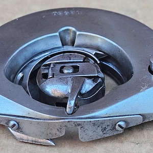 May include: Close-up of a vintage sewing machine bobbin case. The metal component is silver-toned with a circular design, featuring a central mechanism and two screws. The overall appearance suggests a well-used, durable piece of equipment.