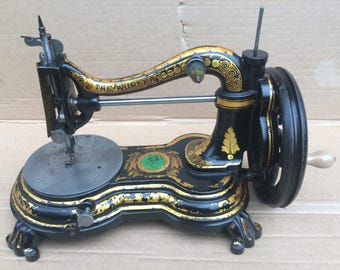 Puede incluir: Máquina de coser antigua negra con detalles dorados y un diseño verde. La máquina tiene una manivela y una placa de aguja.
