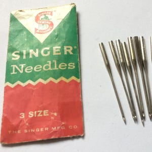 Puede incluir: Un paquete de agujas de máquina de coser Singer vintage con el texto "SINGER Needles" en blanco sobre un fondo rojo y verde. Varias agujas de coser plateadas están esparcidas junto al paquete. El paquete dice "3 SIZE".