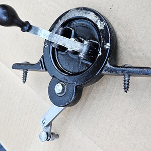 May include: A vintage, cast iron hand-cranked mechanism, painted black, with a wooden handle. The device features a circular gear system, a silver-colored lever, and is mounted with screws. The overall design suggests a historical or industrial tool.