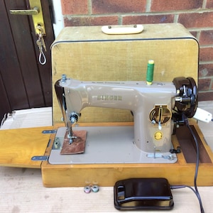 Könnte beinhalten: Eine Vintage-Nähmaschine von Singer in einem Holzkasten mit Fußpedal. Die Maschine ist beige mit einem schwarzen Fußpedal und in gutem Zustand.