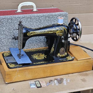 Op de afbeelding: Een vintage zwarte Singer naaimachine met gouden accenten en een houten kist. De machine is in goede staat en wordt geleverd met een voetpedaal en een verscheidenheid aan accessoires.