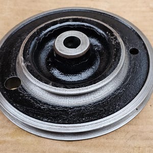 Op de afbeelding: Een cirkelvormige metalen katrol met een zwart midden en een zilverkleurige buitenrand. De katrol heeft een centrale naaf en twee kleine gaten. Waarschijnlijk gebruikt in machines of mechanische toepassingen.