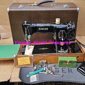 May include: A vintage Singer sewing machine in black with gold lettering, displayed with its brown carrying case and accessories. The machine sits on a wooden base, with a green extension table. A foot pedal is included.