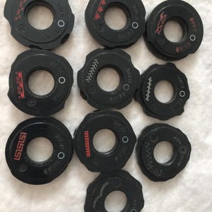 May include: A collection of black sewing machine bobbins with various red and white stitch pattern designs. Each bobbin has a central hole and the text "MADE SWISS".