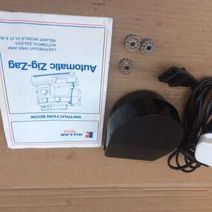 May include: A black Riccar Automatic Zig-Zag sewing machine instruction booklet, a black foot pedal, a white power cord, and three silver bobbins.