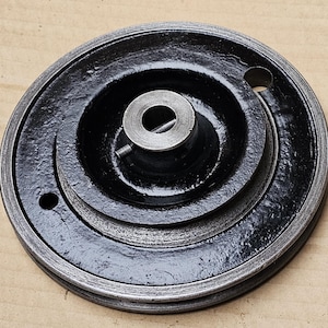 Puede incluir: Polea circular de metal negro con borde y cubo central plateados. La polea tiene un borde exterior acanalado y un orificio central para un eje. Se ven dos pequeños agujeros en la superficie de la polea. Probablemente sea un componente de una máquina.