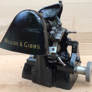 Puede incluir: M&aacute;quina Willcox & Gibbs de metal negro con una cubierta de metal negro y una perilla plateada. La m&aacute;quina tiene una peque&ntilde;a rueda de metal y una palanca.