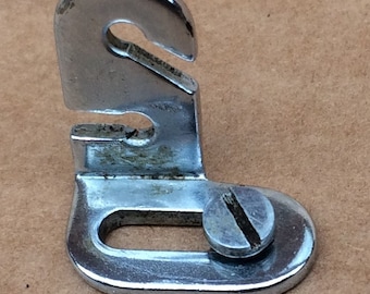 May include: A silver metal sewing machine foot with a hook and a screw. The foot is designed to be attached to a sewing machine.