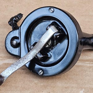 May include: A black metal hand-crank mechanism featuring a circular housing with visible screws, a silver-toned metal arm, and a dark wooden handle. The device includes a protruding side attachment point with a circular hole for mounting purposes.