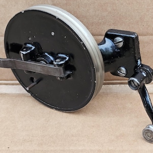 May include: A vintage black and silver mechanical component with a circular disc, a lever, and a handle. The disc has a silver rim and is connected to a black arm with a handle. The component appears to be made of metal.