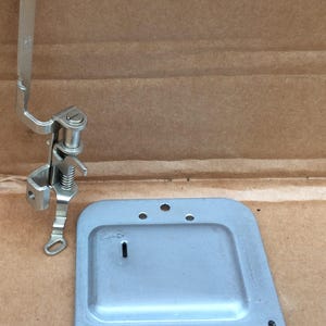 May include: A set of vintage sewing machine parts. The parts are made of silver-colored metal and include a presser foot assembly and a rectangular plate with several holes and a slot. The parts are likely for a vintage sewing machine.