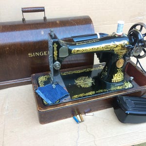 Op de afbeelding: Een vintage zwarte Singer naaimachine met gouden accenten en een houten kist. De machine heeft een voetpedaal en is in goede staat.