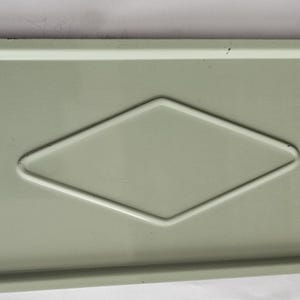 Puede incluir: Una bandeja rectangular de metal verde claro con una hendidura en forma de diamante en el centro. Una llave plateada está sujeta al lado izquierdo. La bandeja tiene un borde ligeramente elevado y una superficie lisa y reflectante.
