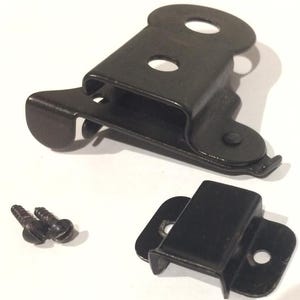 Puede incluir: Un conjunto de componentes de ferretería de metal negro, que incluyen dos soportes, dos tornillos y una pieza articulada con una abertura circular. Las piezas están dispuestas sobre una superficie blanca.