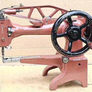 Pode incluir: Máquina de costura industrial vintage em tom castanho-avermelhado. Possui um grande volante preto com cabo de madeira e um conjunto de agulhas. A máquina é projetada para tarefas de costura pesadas.