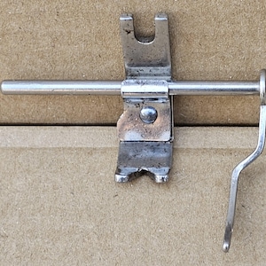 May include: A vintage, silver-toned metal sewing machine part. The piece features a horizontal rod, a U-shaped bracket, and a curved lever. The metal shows signs of wear, with some discoloration and minor imperfections, suggesting age and use.