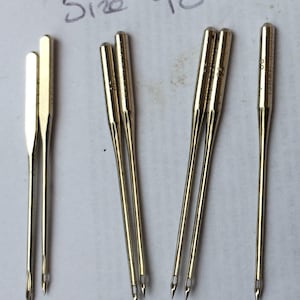Puede incluir: Varias agujas doradas para máquina de coser. Las agujas están dispuestas sobre una superficie blanca. El texto manuscrito "Size 90" en tinta negra es visible encima de las agujas.
