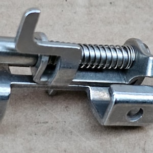 May include: A silver metal sewing machine foot with a spring and a lever. The foot has a curved shape and a hole in the side.