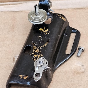 May include: Black sewing machine part with gold floral accents. The part includes a pulley, a bobbin case, and a screw. The metal has some wear and tear, suggesting it is vintage.