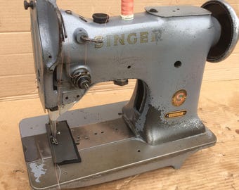 May include: A vintage Singer sewing machine with a gray metal body and a black foot pedal. The machine has a spool of thread on top and a needle in the needle plate. The machine is in good condition, but shows signs of wear and tear.