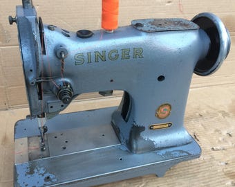 May include: A vintage Singer sewing machine in blue with a spool of orange thread. The machine has a metal base and a metal arm with a needle and thread.
