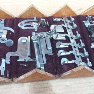 May include: A wooden box with a velvet lining containing a collection of vintage sewing machine feet. The feet are made of metal and have different shapes and sizes. The box is open and the feet are visible.