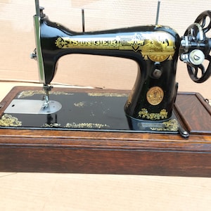 Op de afbeelding: Antiek zwarte Singer naaimachine met gouden accenten en een houten kist. De machine heeft een handwiel en een naald.