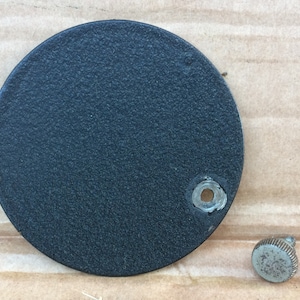 Puede incluir: Una tapa redonda de metal negro con un pequeño agujero en el centro. También se muestra un tornillo de metal plateado.