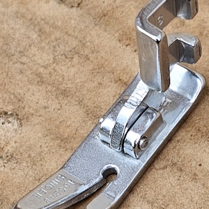 May include: A silver sewing machine foot with a spring-loaded mechanism and the text "1052418 SMOOTH" on the bottom.