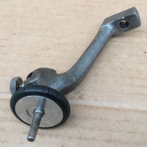 Puede incluir: Un pie de máquina de coser de metal plateado con una rueda de goma negra y una pequeña perilla. El pie está diseñado para ser fijado a una máquina de coser.