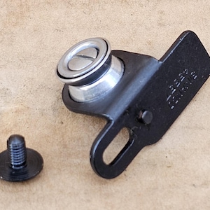 May include: A black metal component with a silver-colored, circular piece and a screw. The component has a rectangular section with stamped text. The screw is separate from the main piece.