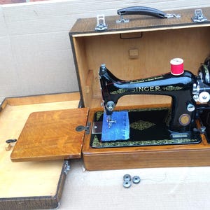 Könnte beinhalten: Eine vintage schwarze Singer Nähmaschine in einem Holzkoffer mit einer blauen Stoffspule und einer roten Garnrolle. Der Koffer hat ein braunes Lederäußeres und ein Holzinnenleben.